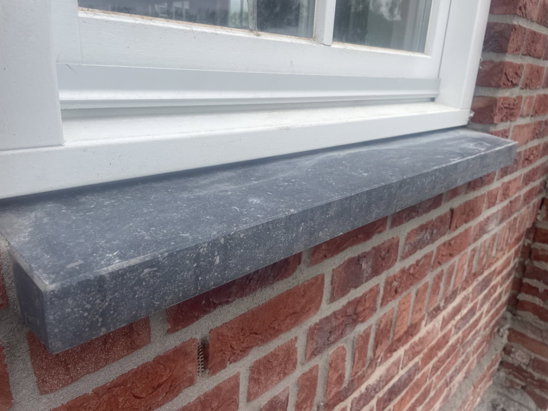 Natuurstenen vensterbank van donkergrijs natuursteen onder een wit raam op een bakstenen muur.